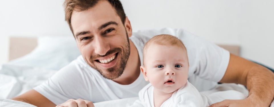 Glücklicher Vater und Baby schauen in die Kamera und liegen auf einem Bett.