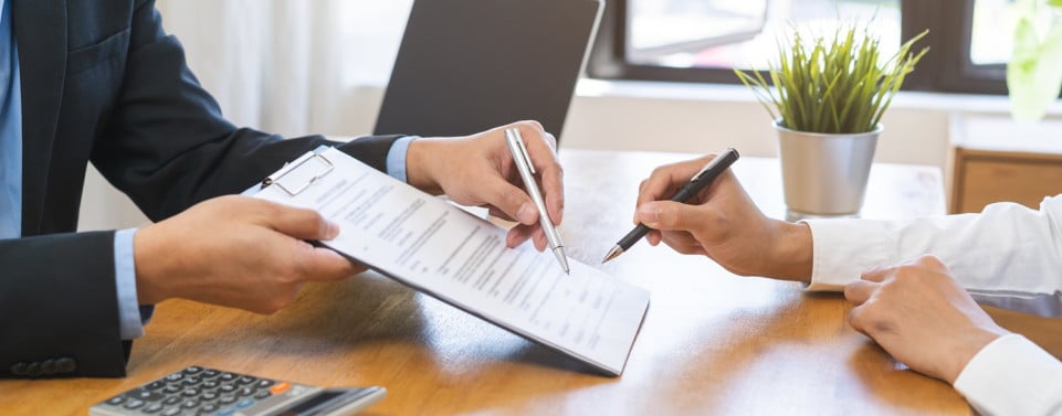 Zwei Personen sitzen sich an einem Schreibtisch gegenüber. Die eine Person hält der anderen ein Dokument zum unterschreiben hin. Die andere Person hält einen Stift und unterschreibt.