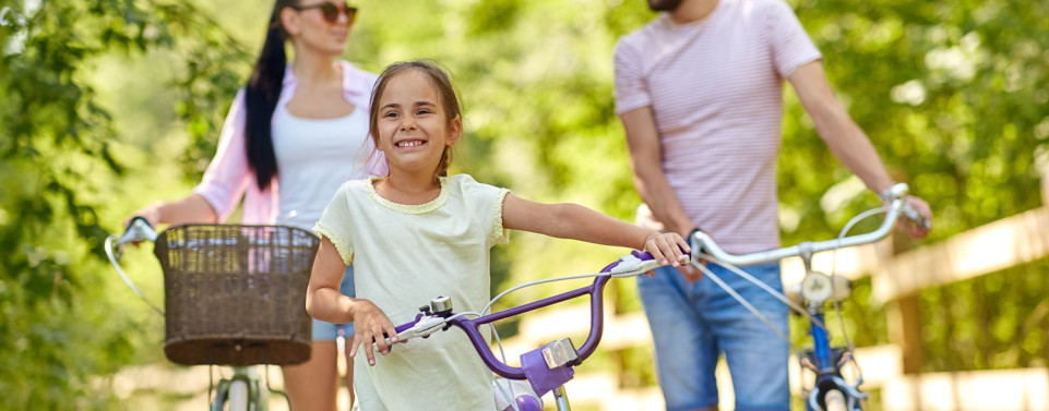 Mädchen schiebt ihr Fahrrad und lacht. Ihre Eltern sind hinter ihr und schieben ihre Räder.