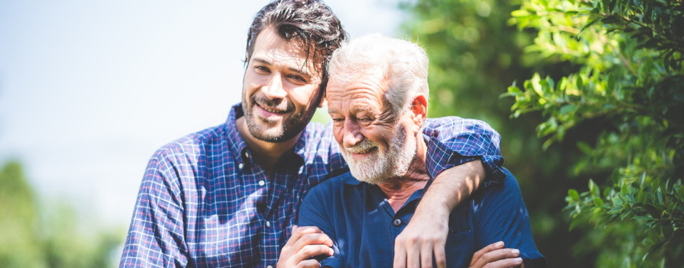 Ein jüngerer Mann hat den Arm um einen älteren Mann gelegt. Beide lächeln.