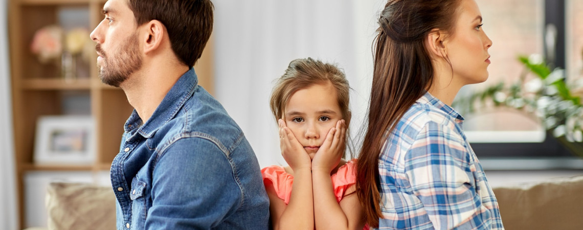 Eltern sitzen mit Kind auf dem Sofa. Kind ist unglücklich und sitzt in der Mitte. Eltern drehen sich den Rücken zu.