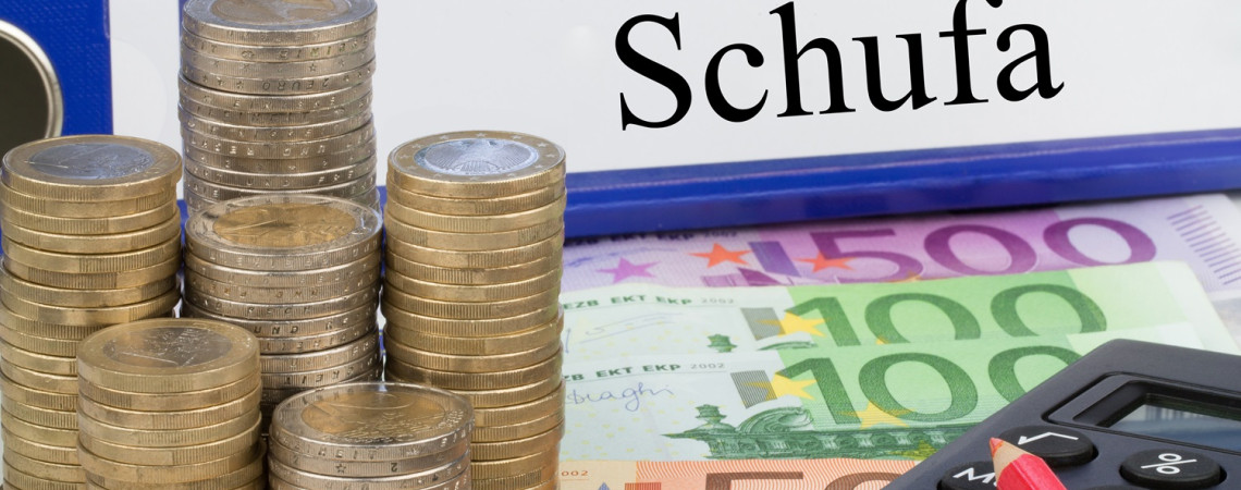 Ein Ordner auf dem "Schufa" steht und davor Geldscheine und Türme aus Euromünzen sowie ein Taschenrechner.