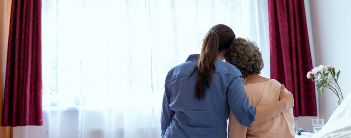 Pflegerin sitzt mit Patientin auf dem Bett und hat den Arm um sie gelegt.