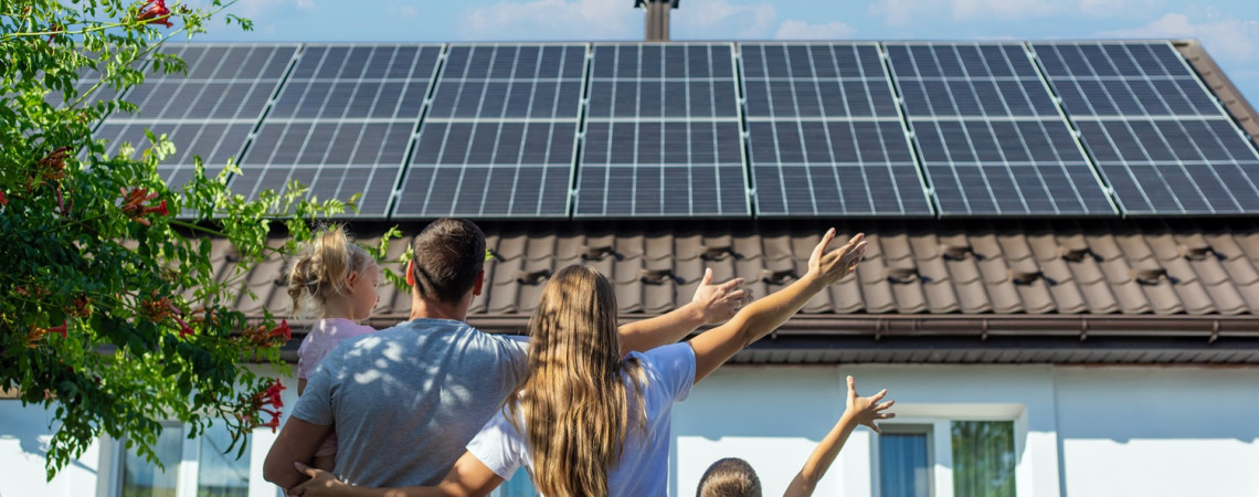 Photovoltaikanlage auf dem Dach eines Hauses. Vor dem Haus steht eine Familie, schaut aufs Dach und hebt freudig die Arme.