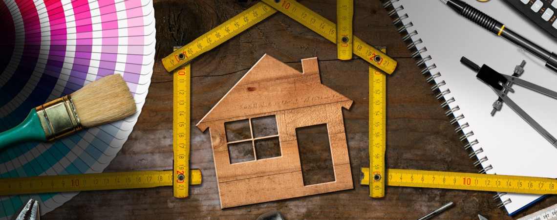 Symbolbild für Umbauten an einem Haus. Auf einem Tisch liegt ein falsches Holzhaus. Ein Zollstock umrahmt das Haus und ist so angeordnet, dass er selbst die Form eines Hauses hat. Es liegen, Pinsel, Hammer, Zirkel und Taschenrechner auf dem Tisch.