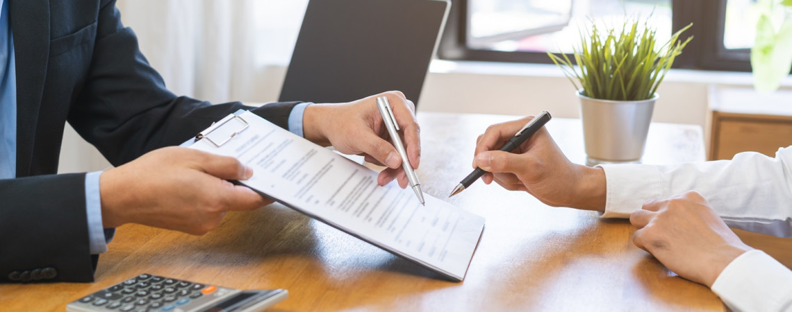 Zwei Personen sitzen sich an einem Schreibtisch gegenüber. Die eine Person hält der anderen ein Dokument zum unterschreiben hin. Die andere Person hält einen Stift und unterschreibt.