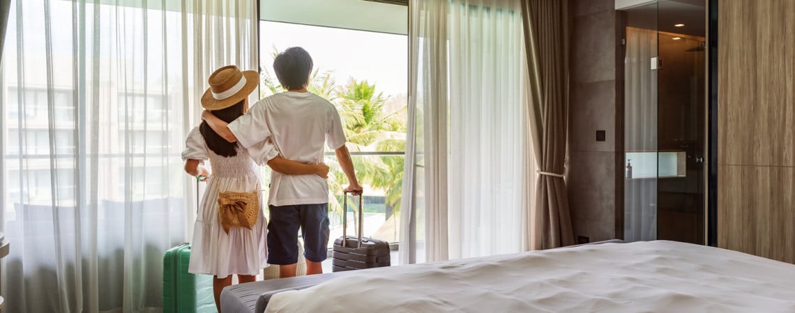 Pärchen steht im Hotelzimmer mit Koffer und schaut aus dem Fenster.
