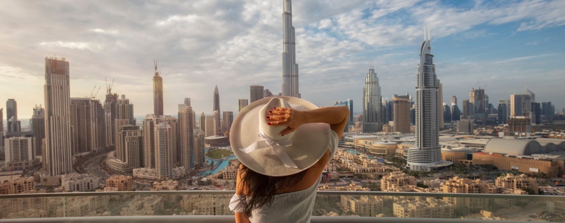 Frau mit Hut steht auf Balkon und blickt auf die Skyline von Dubei.