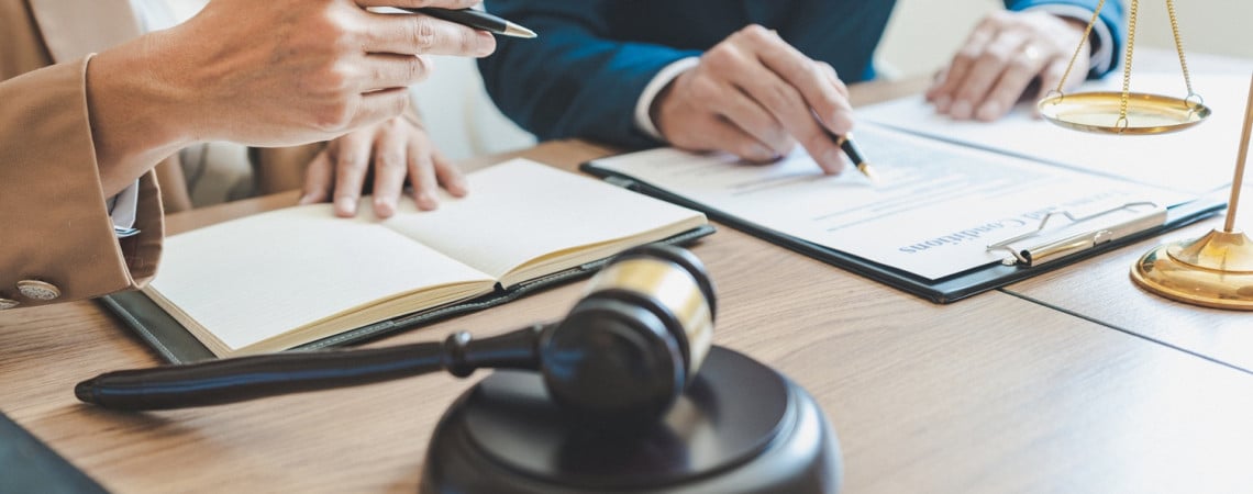 Mann im Anzug am Schreibtisch. Ein Richterhammer auf dem Schreibtisch und eine andere Person, mit einem Stift in der Hand. 