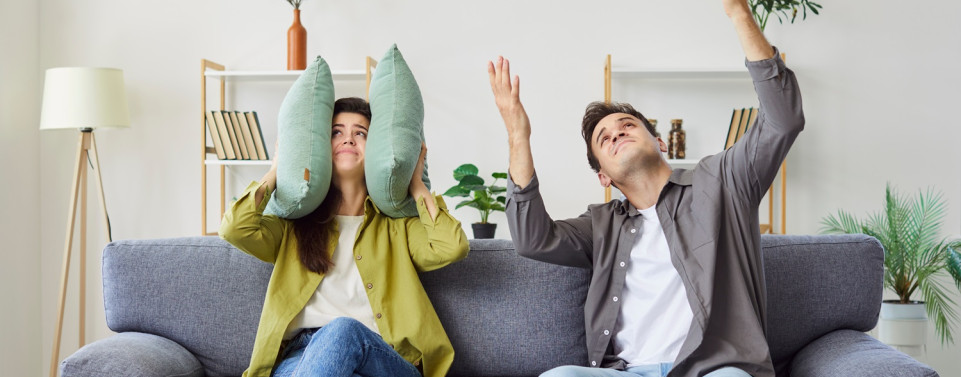 Lärmbelästigung durch Nachbarn. Eine Person sitzt auf einem Sofa und hält sich Kissen auf die Ohren. Ein andere Person sitzt neben ihr und hält die Hände in die Luft.