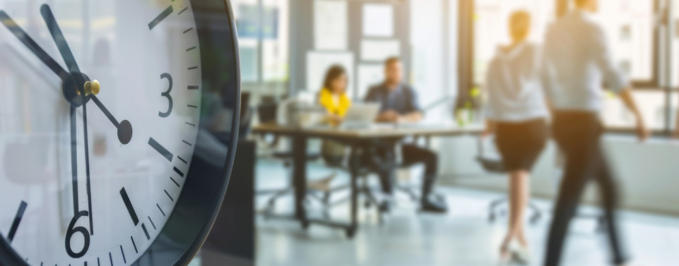 Symbolbild für Arbeitszeitbetrug, indem links ein großer Wecker zu sehen und im Hintergrund unscharf ein Büro mit Menschen.