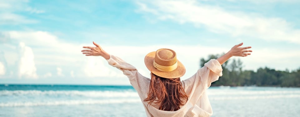 Frau mit Hut hebt die Arme. Sie steht am Meer.