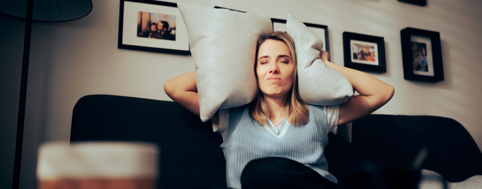 Frau sitzt auf Sofa, hält sich Kissen auf die Ohren und hat einen genervten Gesichtsausdruck.