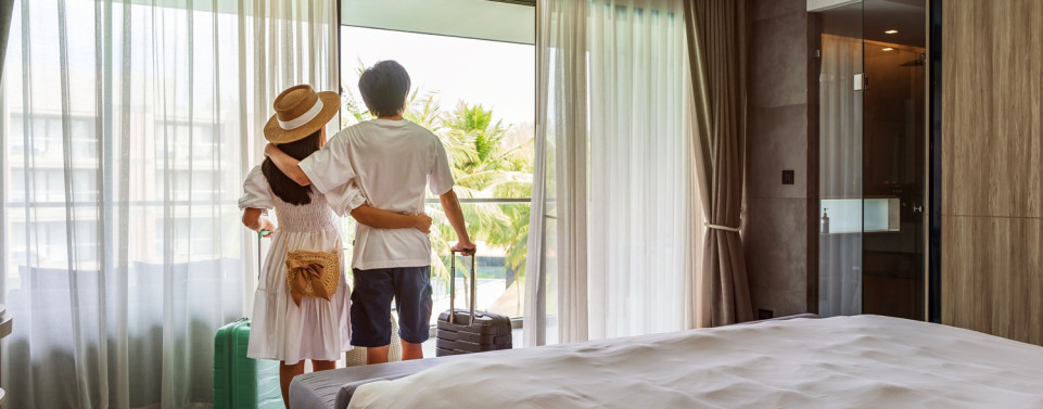 Pärchen steht im Hotelzimmer mit Koffer und schaut aus dem Fenster.