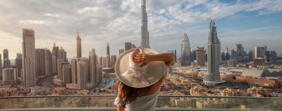 Frau mit Hut steht auf Balkon und blickt auf die Skyline von Dubei.