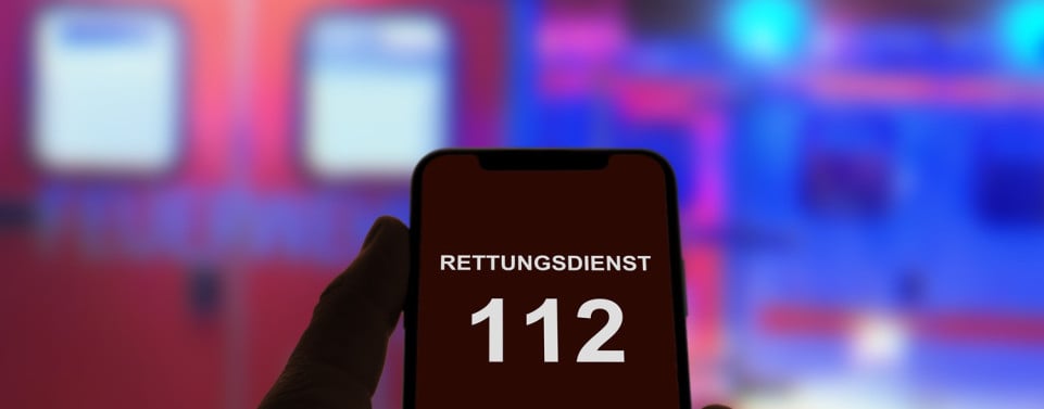 Person hält Smartphone in der Hand und ruft den Notruf 112, im Hintergrund Rettungswagen und Notarzt mit Blaulicht bei Nacht