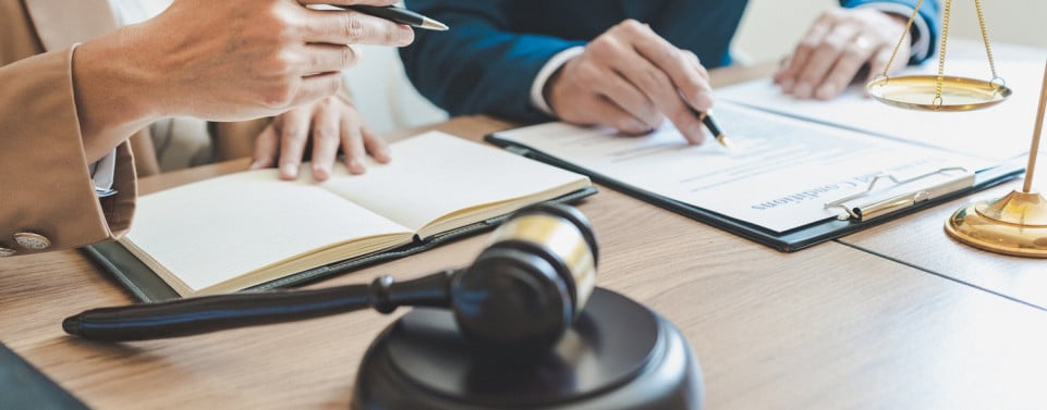 Mann im Anzug am Schreibtisch. Ein Richterhammer auf dem Schreibtisch und eine andere Person, mit einem Stift in der Hand. 