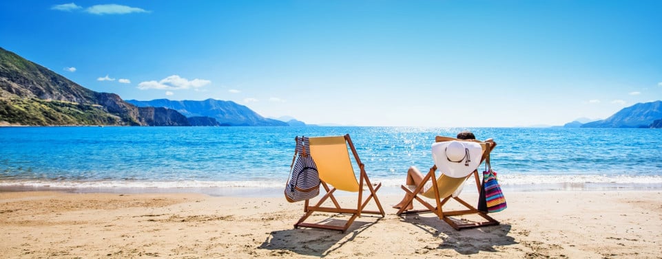 Strand mit Meer, zwei Liegestühle und eine Person mit Sonnenhut in einem der Stühle