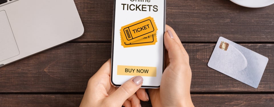 Hände halten ein Smartphone auf dem "Online Tickets" steht und ein "Buy now" Button zu sehen ist.